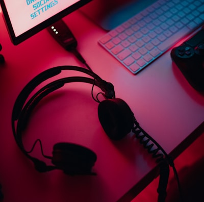 A dimly lit gaming setup with a monitor displaying a game's menu on the screen. A mechanical keyboard sits in front of the monitor. A pair of over-ear headphones and a game controller rest on the desk. The scene is bathed in a red and blue light, creating a moody gaming atmosphere.