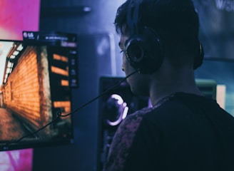 A person wearing headphones is engaged in gaming on a computer, with a large screen displaying a video game scene, showing a wooden building and a path.