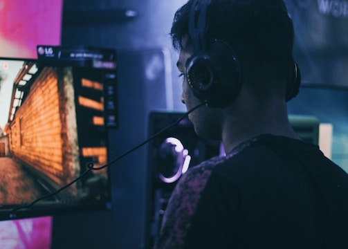 A person wearing headphones is engaged in gaming on a computer, with a large screen displaying a video game scene, showing a wooden building and a path.