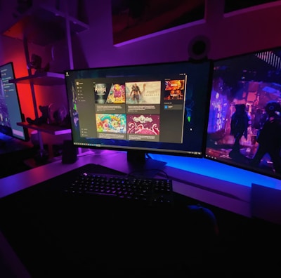 A gaming setup featuring multiple screens with vibrant game-related images illuminated by colorful LED lighting. The environment is dimly lit, highlighting the vivid displays on the monitors. A keyboard is visible on the desk beneath the central monitor.