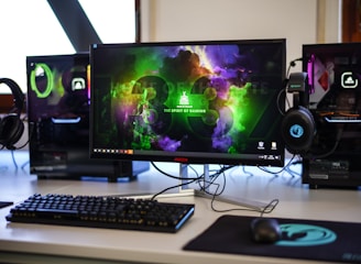 A gaming setup featuring a central monitor with a vibrant display showing 'The Spirit of Gaming'. On either side, there are gaming PCs with illuminated components and transparent cases. Headsets hang from the side of the monitor. The desk also has a black keyboard and mouse, and a branded mousepad.