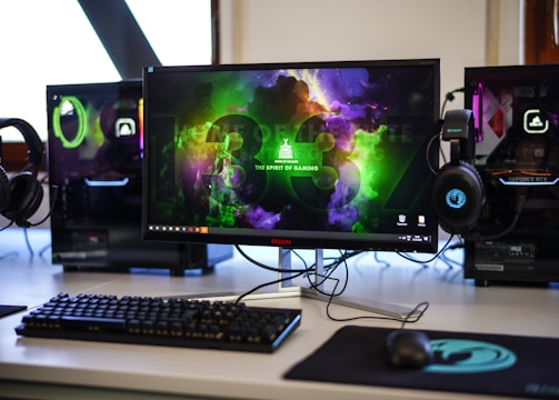 A gaming setup featuring a central monitor with a vibrant display showing 'The Spirit of Gaming'. On either side, there are gaming PCs with illuminated components and transparent cases. Headsets hang from the side of the monitor. The desk also has a black keyboard and mouse, and a branded mousepad.