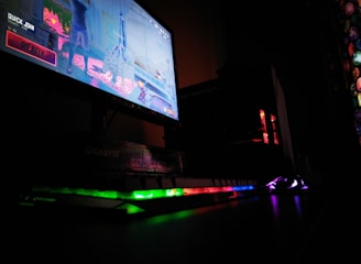 A dimly lit gaming setup is highlighted with a computer monitor displaying a game screen. The surrounding area is dark, with RGB lighting from a keyboard and PC tower creating vibrant reflections on the desk.