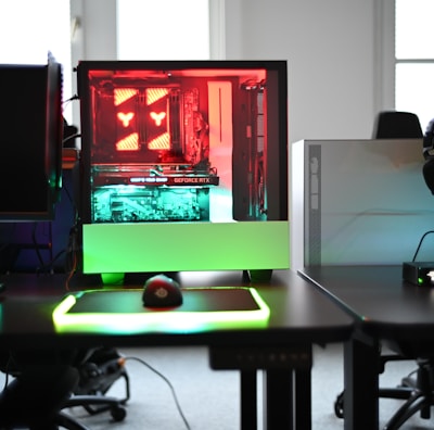 A modern gaming setup is displayed on a desk with multiple computer monitors and an open gaming PC tower. The tower is illuminated with red and green LED lights, showcasing a 'GeForce RTX' graphics card. A gaming mouse rests on a mousepad with glowing edges, and there is a keyboard with vibrant backlighting. The room appears well-lit with visible windows in the background.