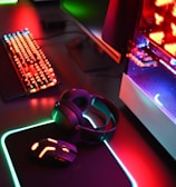 A gaming setup with a mechanical keyboard featuring multicolor RGB lighting, a glowing mouse on a mousepad with illuminated edges, a set of headphones, and a high-end gaming PC with a transparent side panel showcasing internal components and red LED lights.