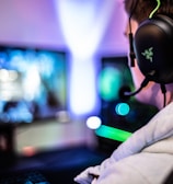 A person wearing headphones is sitting in front of a computer setup with multiple monitors displaying colorful, blurred graphics. The headphones have a distinctive logo illuminated in green. The ambient lighting and screen glow add to the immersive gaming environment.