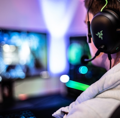 A person wearing headphones is sitting in front of a computer setup with multiple monitors displaying colorful, blurred graphics. The headphones have a distinctive logo illuminated in green. The ambient lighting and screen glow add to the immersive gaming environment.