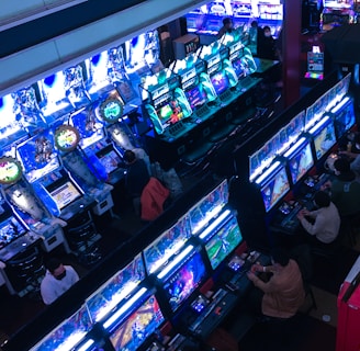 A bustling arcade filled with rows of gaming machines, each with vibrant displays and controls. Several people are seated at these machines, deeply engaged in playing games. Bright fluorescent lights cast a blue and neon glow across the room, enhancing the lively and energetic atmosphere.