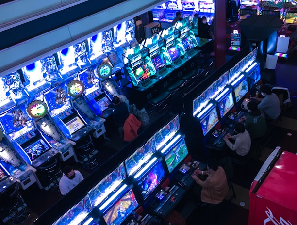 A bustling arcade filled with rows of gaming machines, each with vibrant displays and controls. Several people are seated at these machines, deeply engaged in playing games. Bright fluorescent lights cast a blue and neon glow across the room, enhancing the lively and energetic atmosphere.